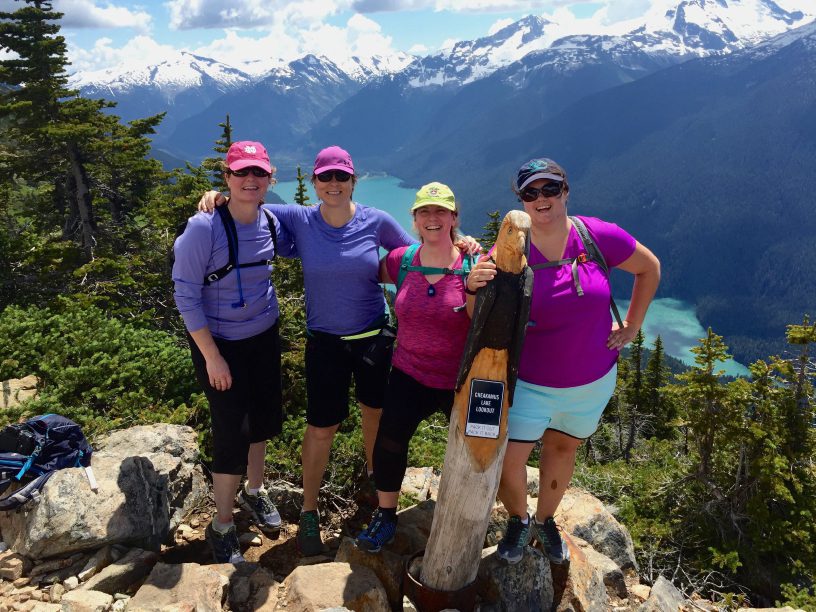 four happy hikers wearing purple overlooking alpine lake - Vifit Vacations
